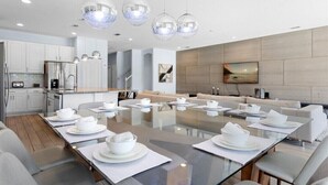 A modern dining area with a glass table set for six, white chairs, and a minimalist kitchen in the background. - Last Minute Disney Getaway! Luxury Villa with Private Pool. Book Now! #66147 (Orlando)