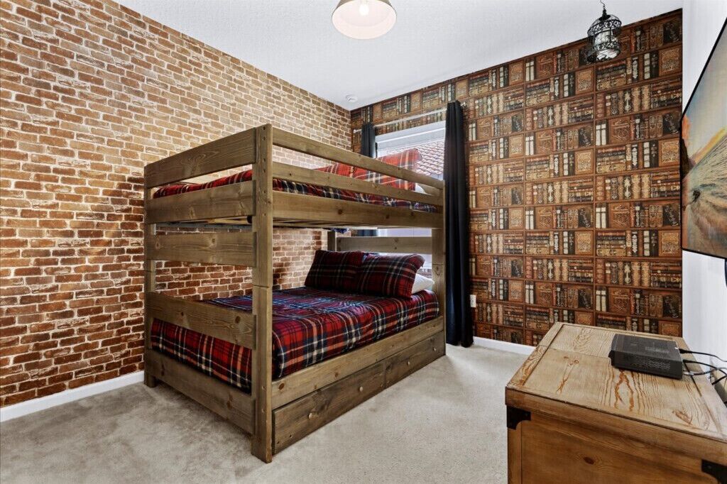 A bunk bed with a plaid blanket, a brick wall, and a bookshelf with books.