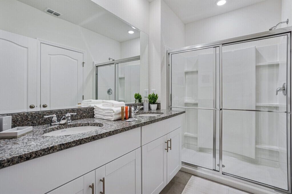 Une salle de bain moderne avec un comptoir en granit, des meubles blancs, un grand miroir, une cabine de douche en verre et une petite plante en pot.