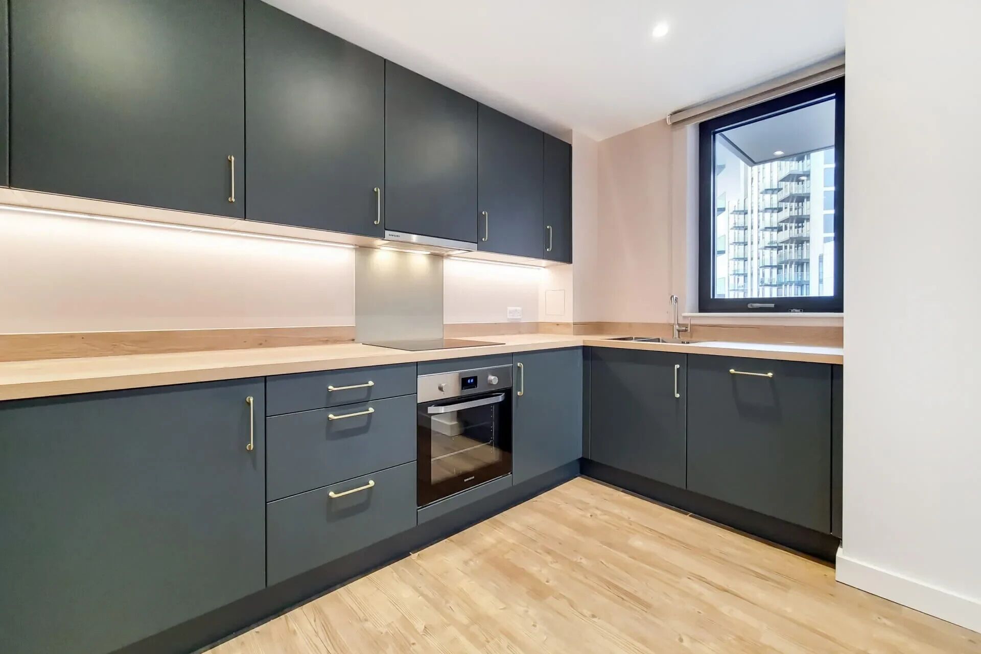 A modern kitchen with dark cabinetry, a built-in oven, and a window with a view of a building.