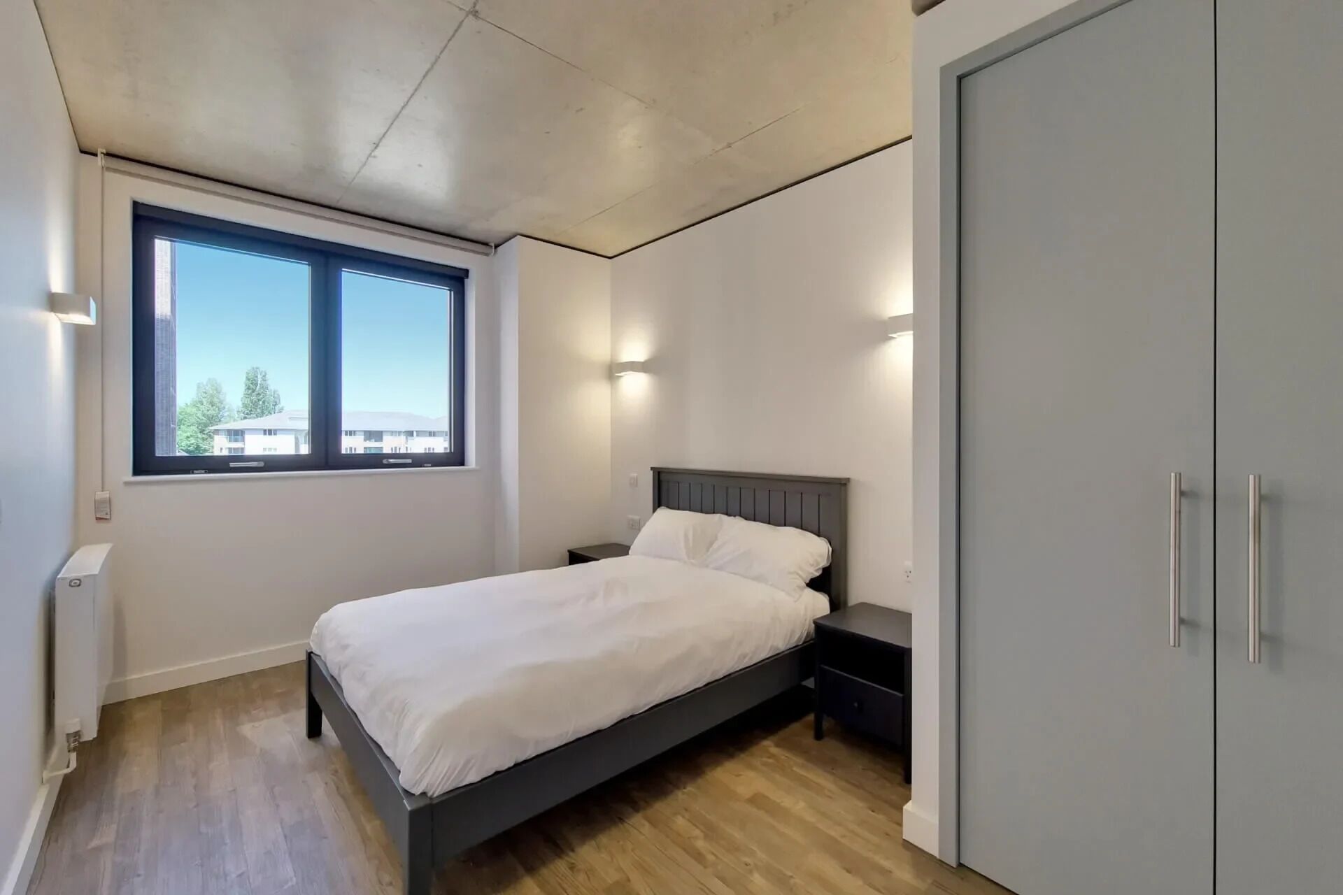 A bedroom with a bed, a nightstand, a wardrobe, and a window with a view of buildings.