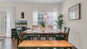 A dining area with a wooden table set for four, surrounded by chairs, a television, and a large window with curtains. - Last Minute Disney Getaway! Private Pool & Themed Bedrooms. Book Now! #65673 (Orlando)