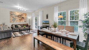 A modern living room with a sofa, coffee table, dining table set for four, and a TV. - Last Minute Disney Getaway! Private Pool & Themed Bedrooms. Book Now! #65673 (Orlando)