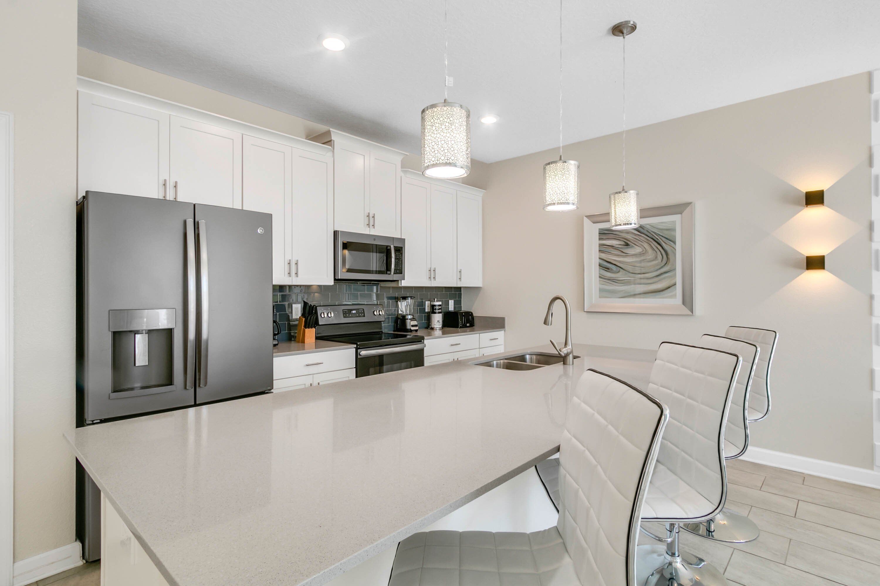 A modern kitchen with white cabinetry, stainless steel appliances, a large island, pendant lighting, and a marble backsplash.