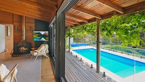 Et moderne udendørs opholdsområde med træterrasse, pejs, swimmingpool og udsigt til et bjergrigt landskab.
