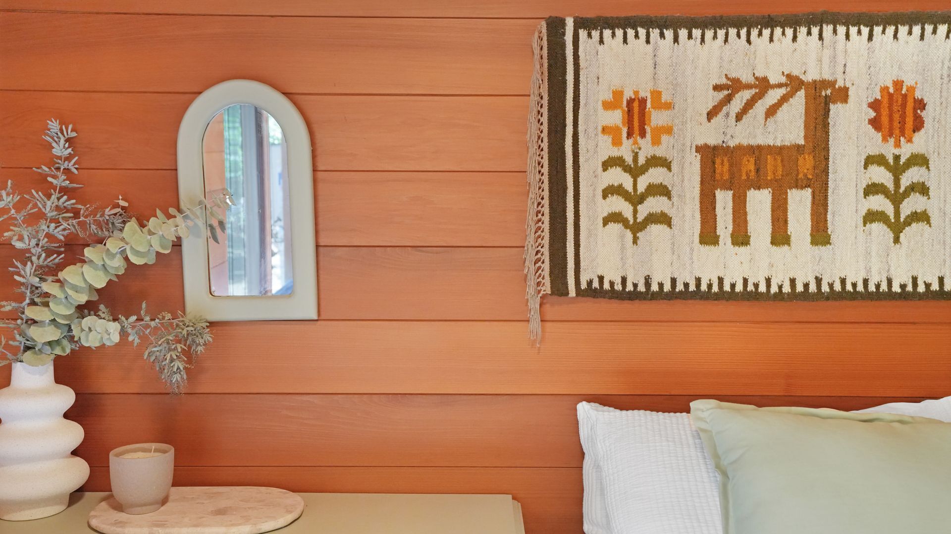 A room with wooden walls, a mirror, a woven tapestry, a vase with plants, and a bed with pillows.