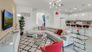 A modern living room with a sofa, coffee table, and red chair, a kitchen area with white cabinets, and a dining table with stools. - Last Minute Disney! Private Pool, Hot Tub, Game Room & Themed Bedrooms. Book Now! #65969 (Orlando)