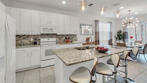 A modern kitchen with white cabinets, granite countertops, and stainless steel appliances. There are bar stools and a dining area with a glass table. - Last Minute Disney! Private Pool, Hot Tub, Game Room & Themed Bedrooms. Book Now! #65969 (Orlando)