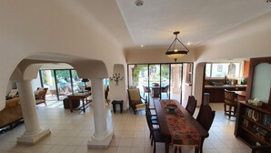 A spacious dining area with a long table, chairs, and a patterned runner. There are columns supporting the ceiling, and a kitchen area is visible in the background.