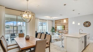 A modern kitchen with a central island, a dining area with a wooden table and chairs, and a view of the outdoors through large windows. - Last Minute Disney Getaway! Private Pool & Themed Bedrooms. Book Now! #65997 (Orlando)