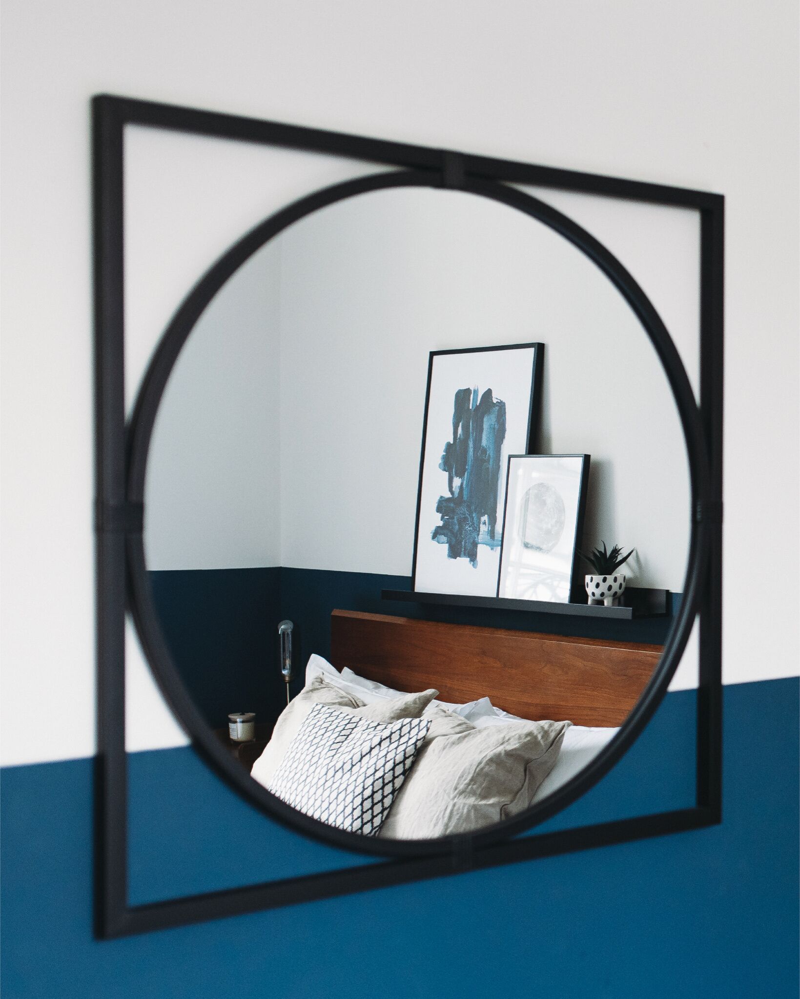 A mirror with a black frame reflecting a room with a blue and white wall, a wooden headboard, and a small shelf with a plant and framed pictures.