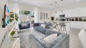 A modern living room with a grey sofa set, a coffee table, and a TV. There is a kitchen area with white cabinets and a dining area with a bar stool. - 5 BD/5BA Themed House Near Disney (Kissimmee)