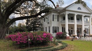 A large, white colonial-style house with a prominent porch and columns, surrounded by lush greenery and flowering bushes.