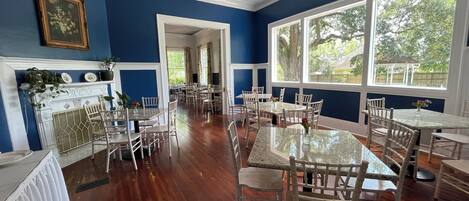 A dining area with multiple tables and chairs, a fireplace, and large windows with a view of greenery.