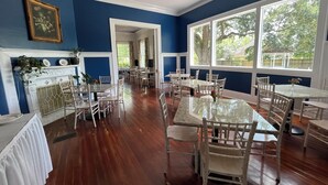 A dining area with multiple tables and chairs, a fireplace, and large windows with a view of greenery. - The Marie Antoinette (Hammond)