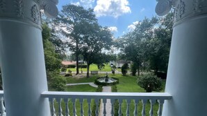 A balcony with white balustrades overlooking a well-maintained lawn with a fountain, surrounded by trees and a clear sky. - The Marie Antoinette (Hammond)