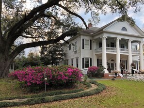 Une grande maison de style colonial, peinte en blanc, dotée d’un porche et de colonnes imposants, et entourée d’une végétation luxuriante et d’arbustes en fleurs.