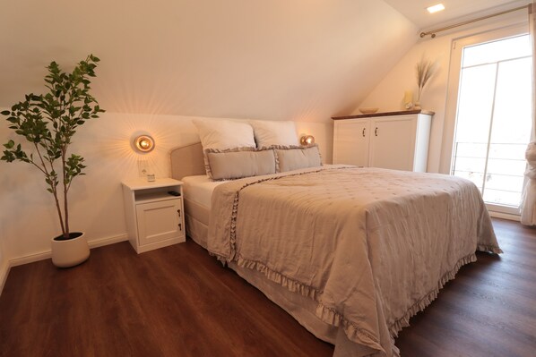 A bedroom with a large bed, bedside tables, a nightstand, a potted plant, and a window with curtains.