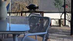 A covered outdoor seating area with a table and chairs, a potted plant, and a view of a building and trees in the background.