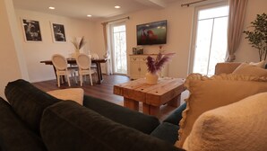 A living room with a dark green sofa, a wooden coffee table with decorative items, a dining area with a white table and chairs, and a TV mounted on the wall.