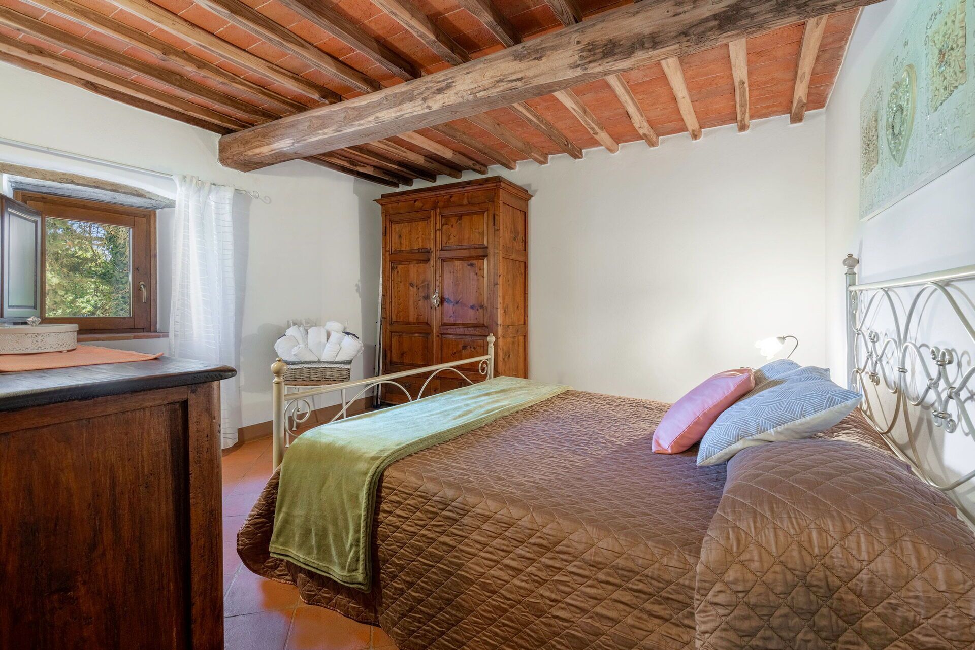 A bedroom with a wooden ceiling, a bed with a quilted cover, a nightstand, a wooden wardrobe, and a window with curtains.