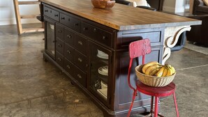 Una isla de cocina con encimera de madera, una silla roja con una cesta de plátanos y un mueble de madera oscura con varios cajones.