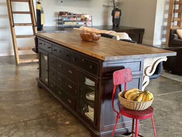 Una isla de cocina con encimera de madera, una silla roja con una cesta de plátanos y un mueble de madera oscura con varios cajones.
