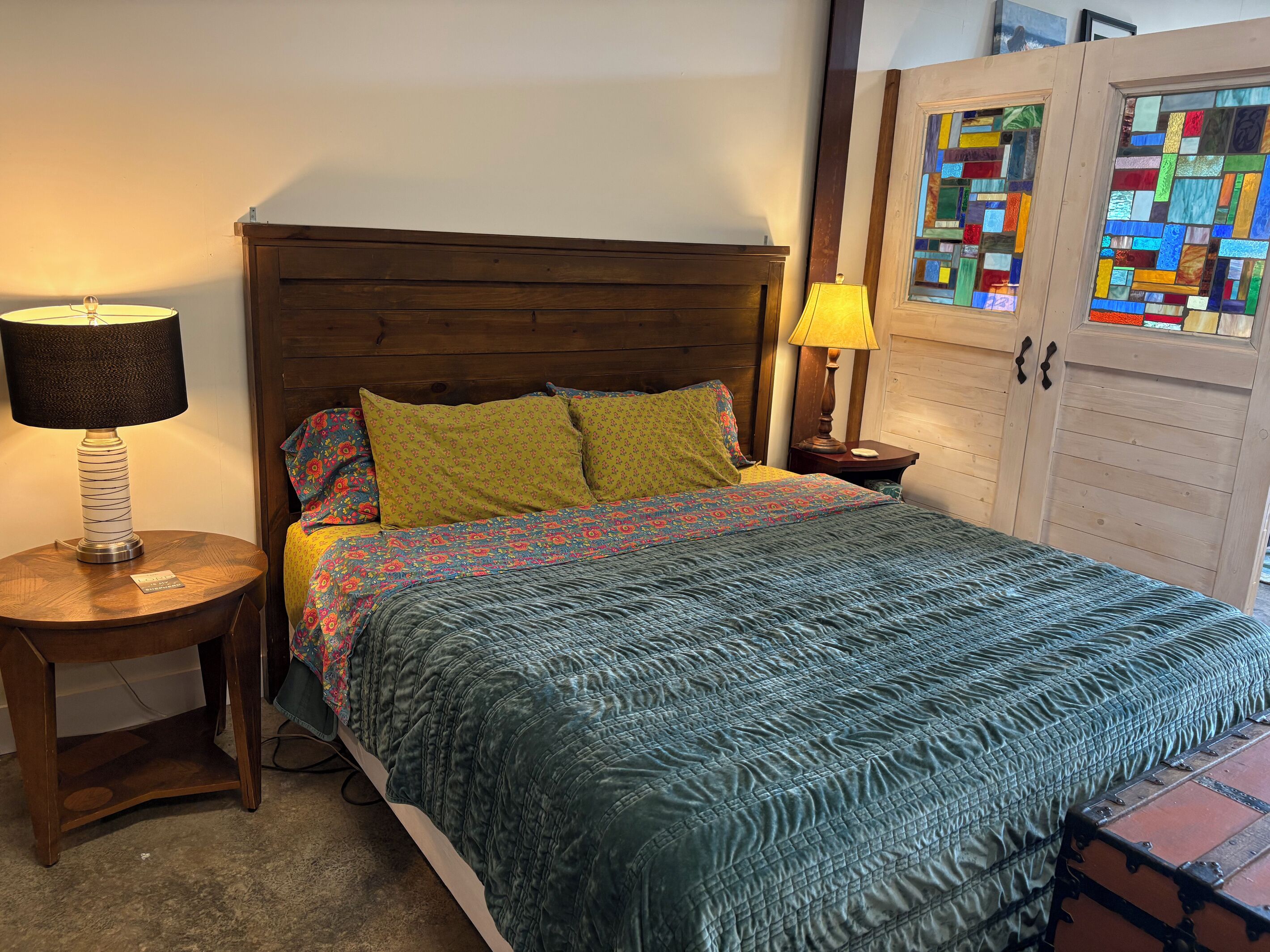 Un dormitorio con cabecera de madera, cama con ropa de cama estampada, mesita de noche con lámpara y un armario con un vitral.