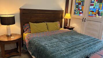 Un dormitorio con cabecera de madera, cama con ropa de cama estampada, mesita de noche con lámpara y un armario con un vitral.