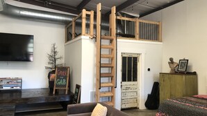 Una habitación estilo loft con una escalera de madera que lleva a un entrepiso, un televisor de pantalla plana, un sofá y una guitarra.