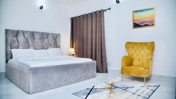 Ein Schlafzimmer mit einem Bett, einem gelben Sessel, einem Gemälde und einem Vorhang.