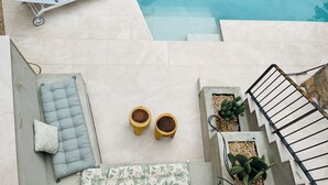 Un área junto a la piscina con una zona de estar acolchada, dos plantas en macetas y una escalera de metal.