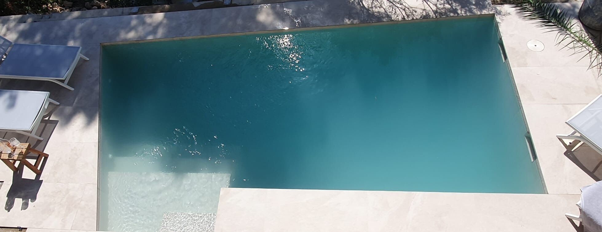 A rectangular swimming pool with a clear blue water surface, surrounded by a tiled deck and shaded by palm trees.