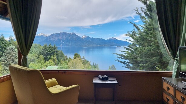 Ein Zimmer mit Blick auf einen See und Berge. Ein Sessel und ein kleiner Tisch mit Fernglas sind vorhanden.