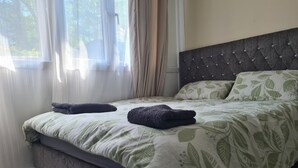 Un lit bien fait, recouvert d’une couvre-lit à motifs de feuilles, avec une tête de lit sombre et deux serviettes pliées posées sur le lit.