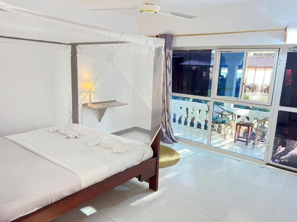 A room with a four-poster bed, a ceiling fan, a window with a view of a balcony, and a small table with a lamp.