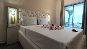 Een hotelkamer met een groot bed, een hoofdbord met een geometrisch patroon, een raam met uitzicht op zee en een wandlamp.