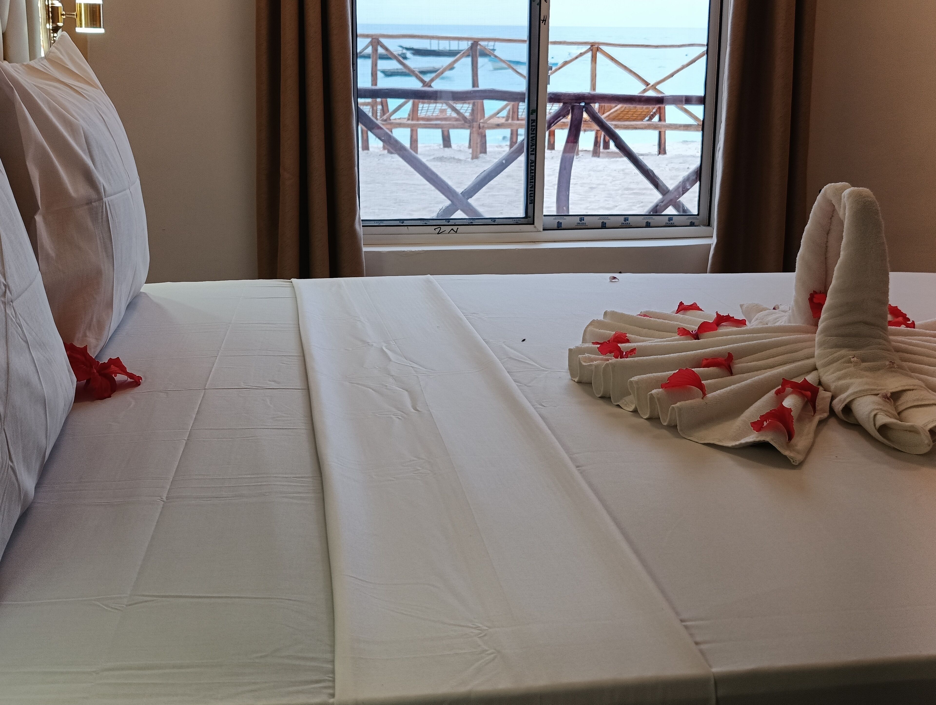 A hotel room with a bed, a window with a view of a beach and wooden pier, and a neatly made bed with towels and red flowers.