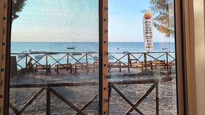 Uitzicht op een strand door een raam, met een houten terras en een metalen hekwerk.