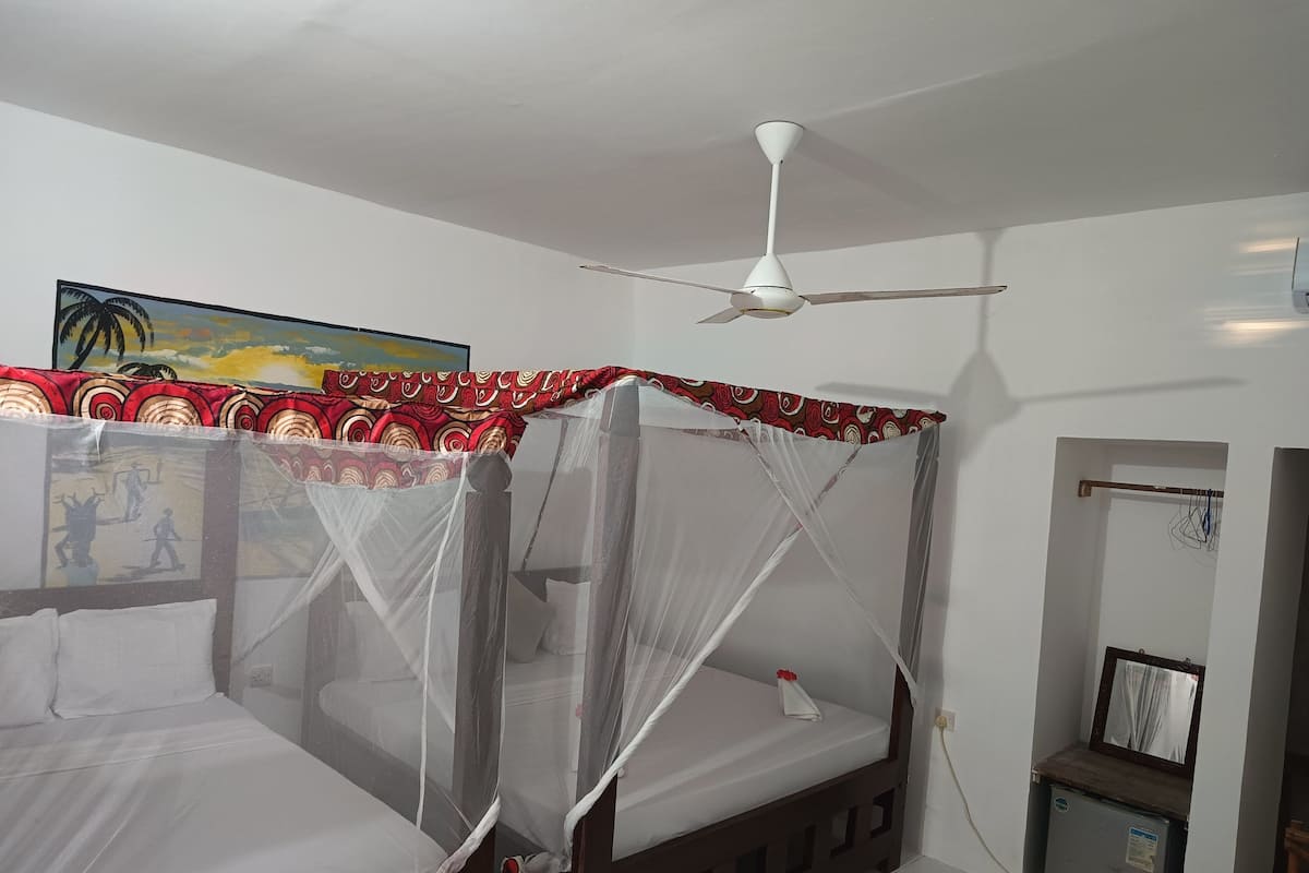 Un dormitori amb un llit sota una mosquitera, un ventilador de sostre i una petita taula de nit.