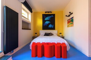 Un dormitorio con una cama, un cuadro de un pez, un cuadro abstracto colorido y un radiador negro.