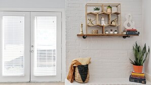Een kamer met een witte bakstenen muur, een houten plank met decoratieve objecten, een deur met jaloezieën en een potplant.
