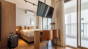 Ein Hotelzimmer mit einem Bett, einem Schreibtisch mit Stühlen, einem Fernseher, einem Koffer und einem Balkon mit Geländer.