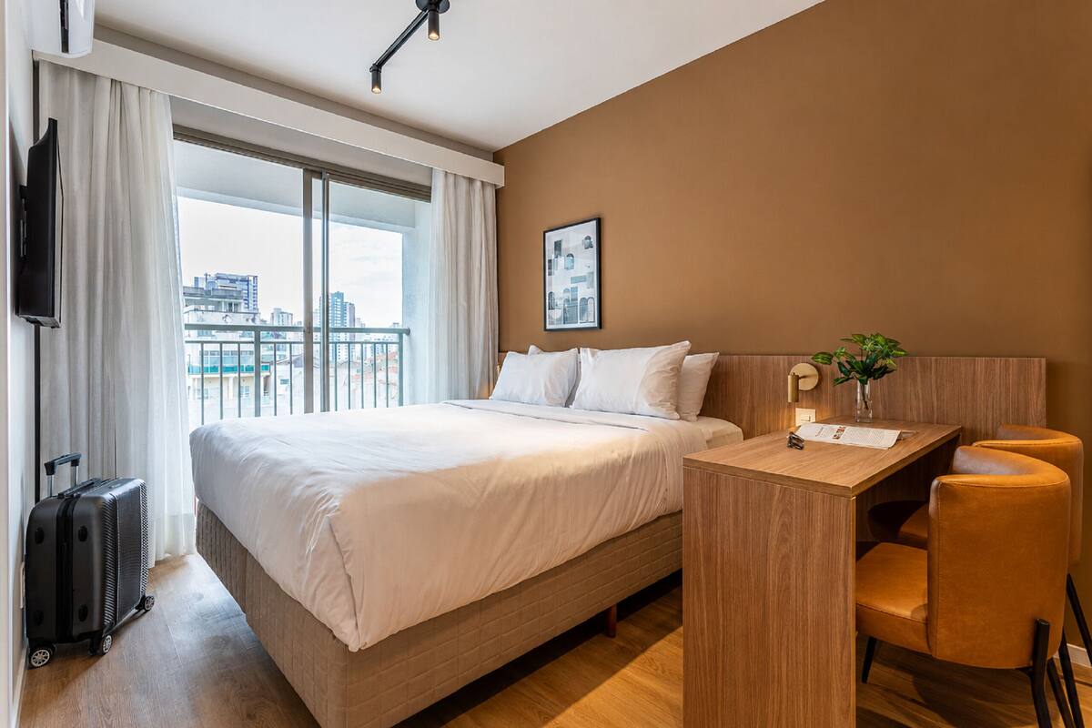Une chambre d’hôtel comprenant un lit, un bureau, une chaise, une télévision, une fenêtre avec des rideaux et une vue sur des immeubles.