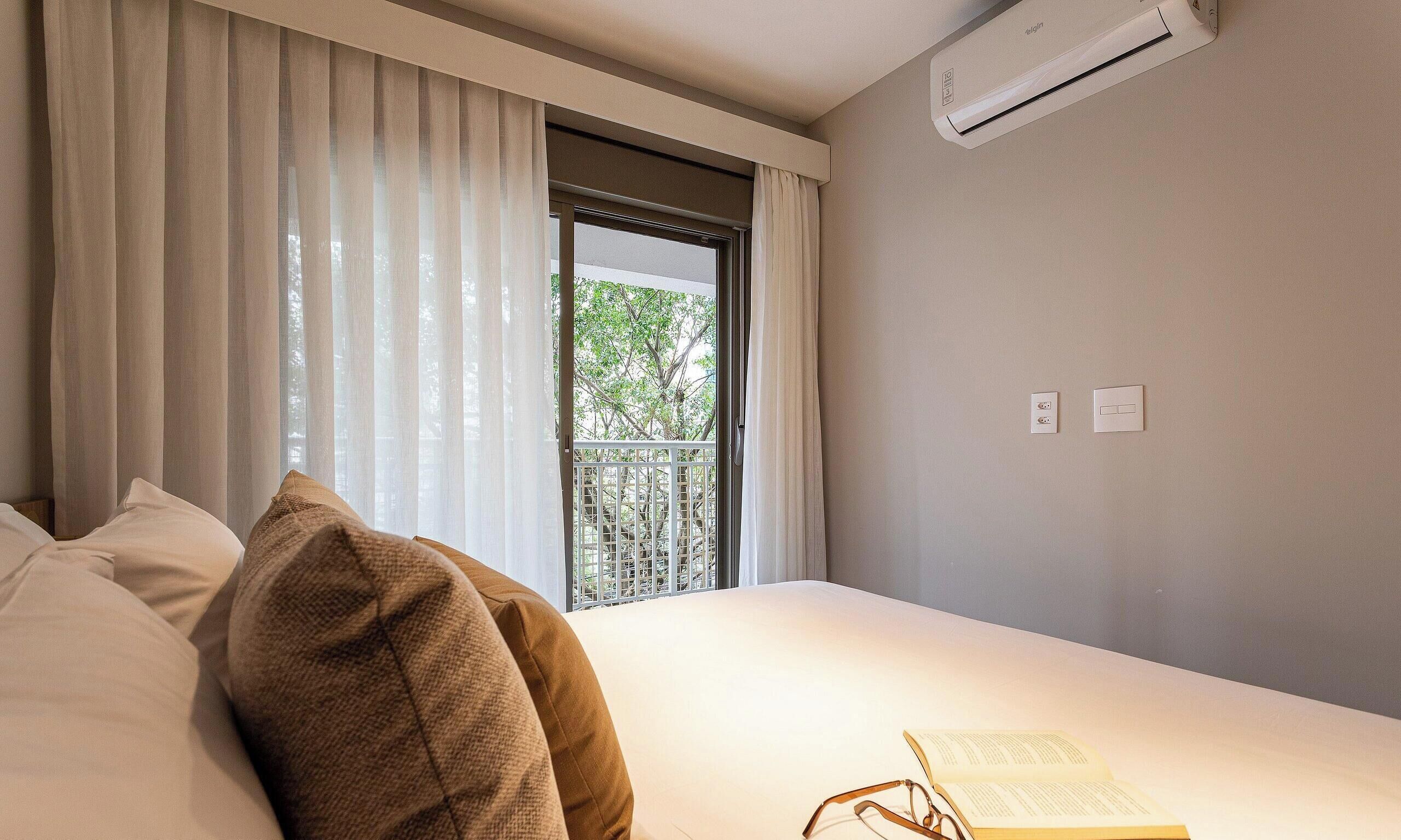 A hotel room with a bed, a window with curtains, an air conditioning unit, and a view of greenery outside.
