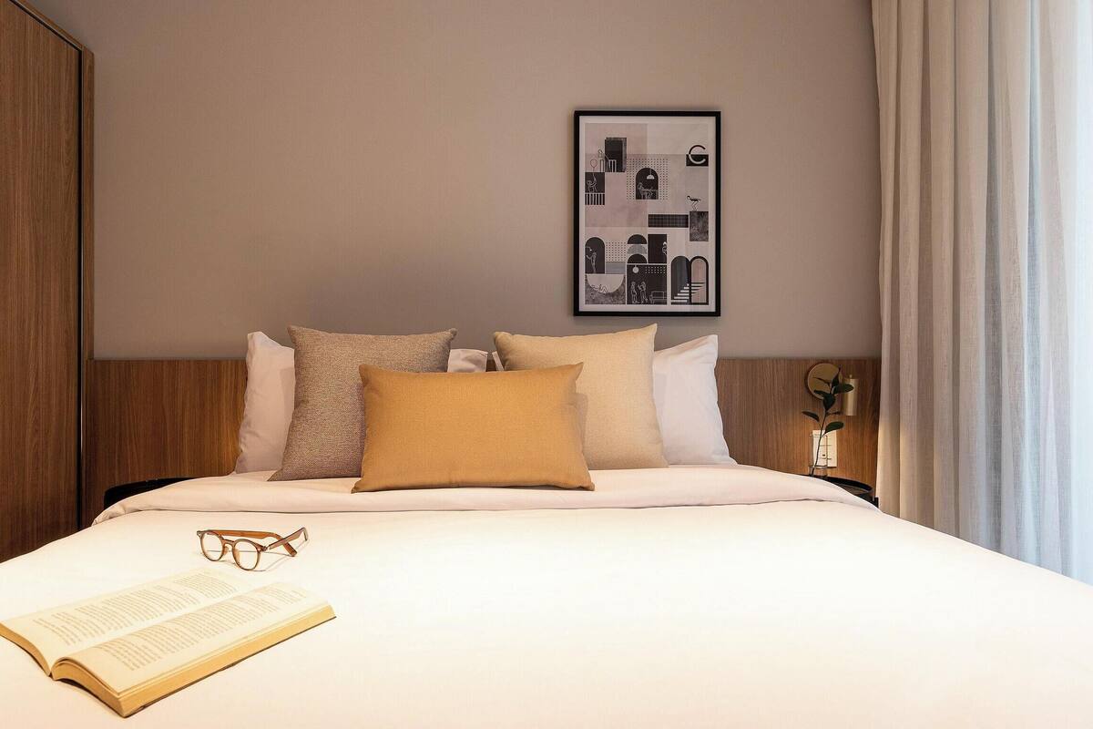 Un lit bien fait, avec des oreillers, un tableau encadré accroché au mur, et un livre et des lunettes posés sur le lit.