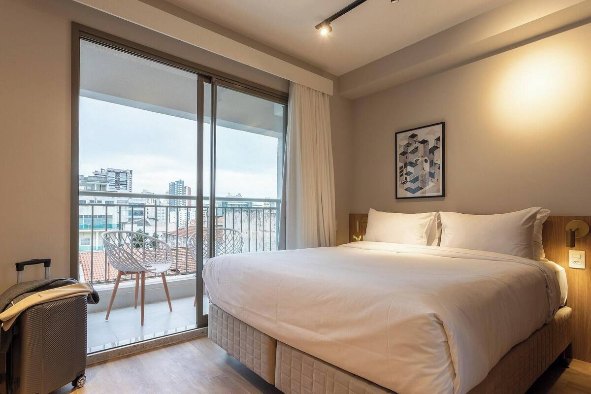 Une chambre d’hôtel avec un grand lit, une vue sur la ville grâce à une porte-fenêtre coulissante, une chaise et une valise.