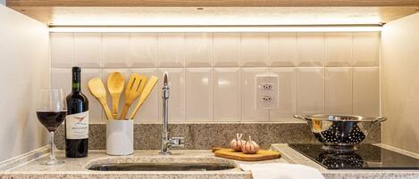 A kitchen with a granite countertop, a sink, a wine bottle, wooden utensils, and a colander on an induction cooktop.