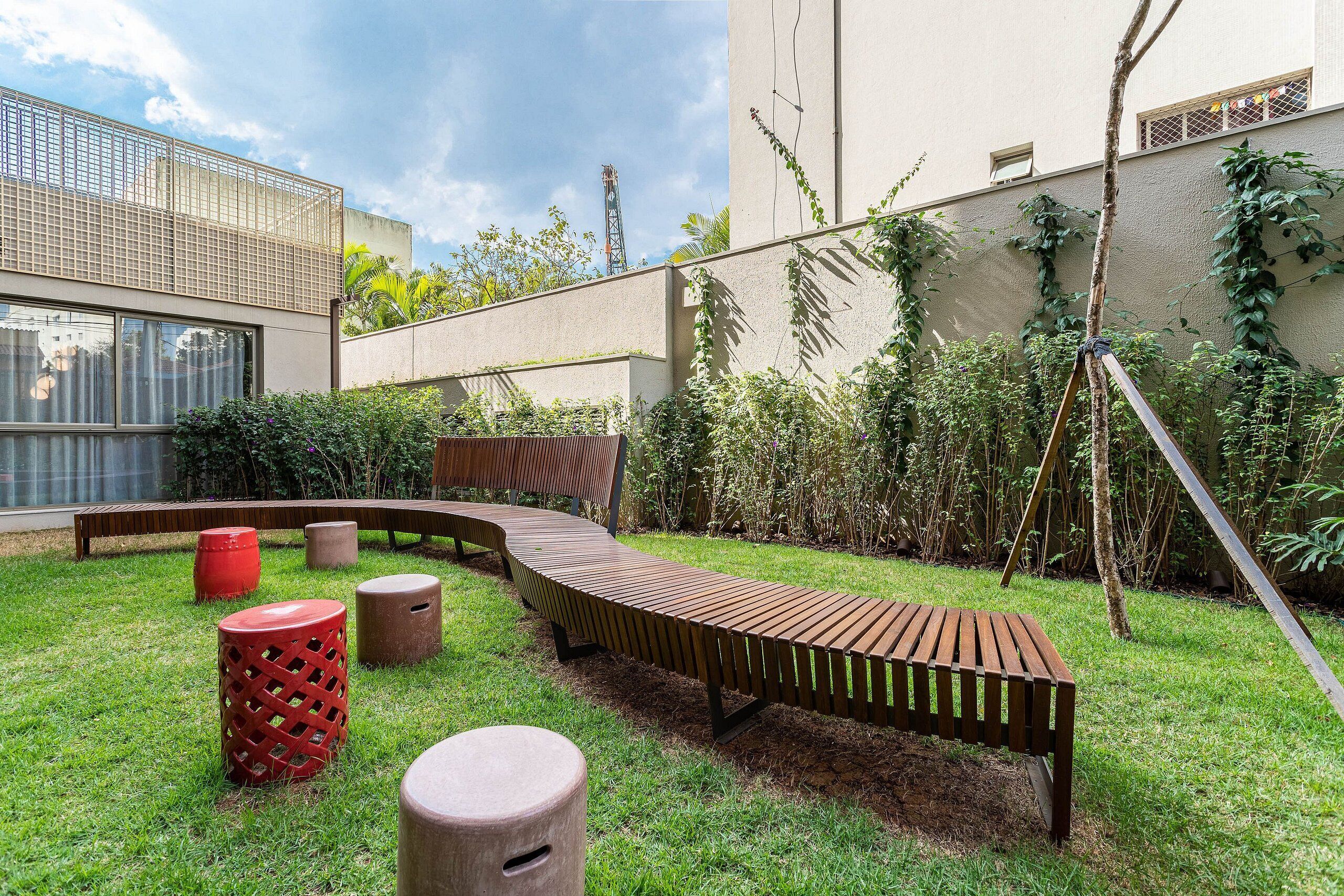 A modern outdoor seating area with a curved wooden bench, red and beige stools, and a swing set.