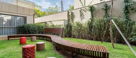 A modern outdoor seating area with a curved wooden bench, red and beige stools, and a swing set.
